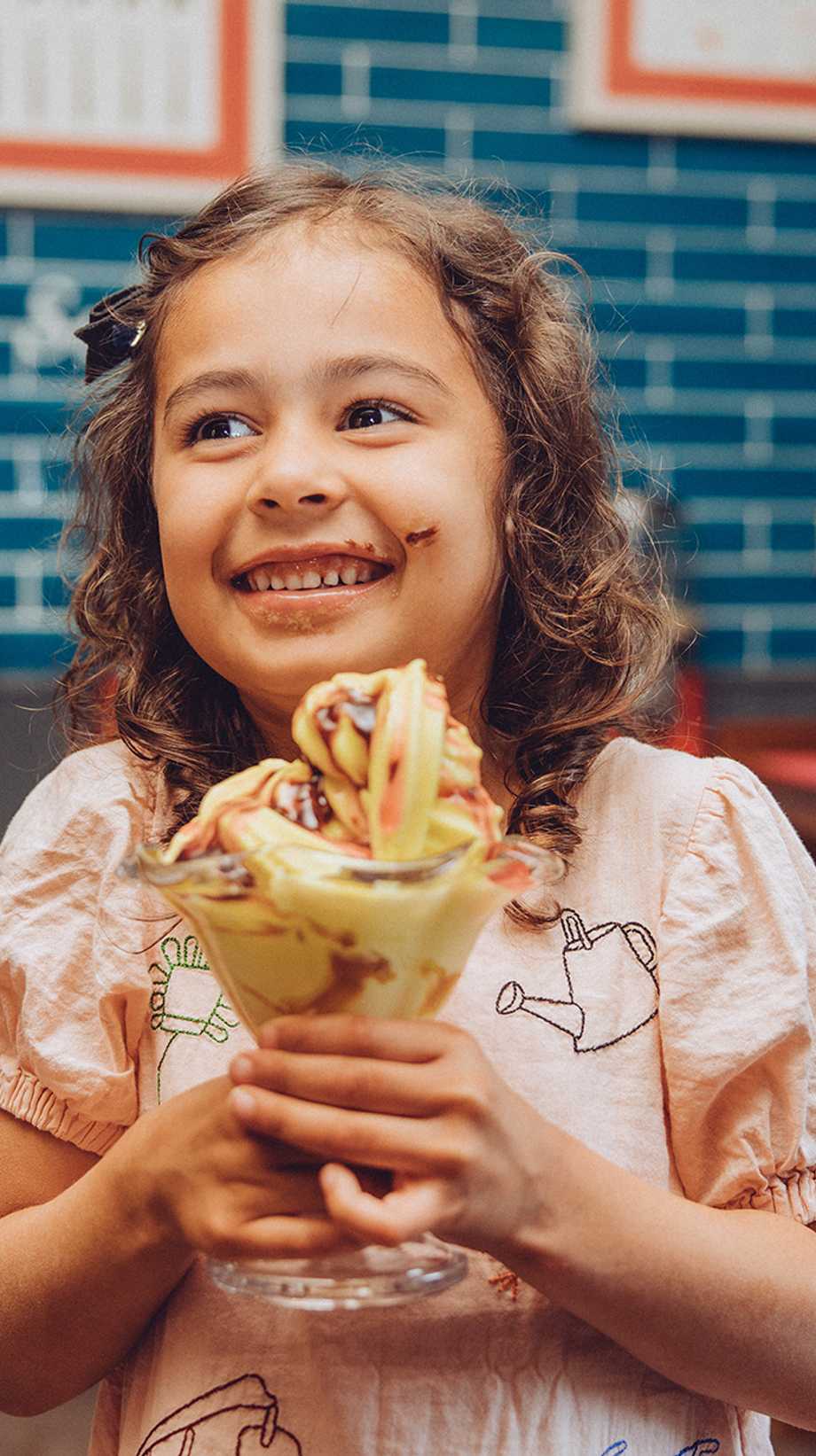 A young girl with chocolate on her cheek is smiling in front of an ice-cream sundae.