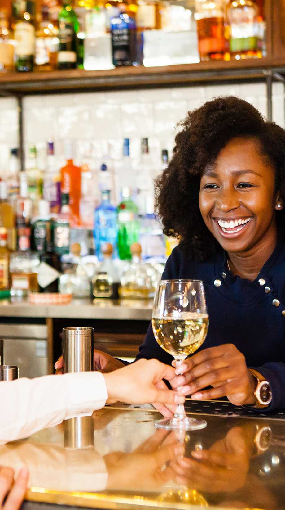 A team member hands a glass of white wine to a guest while smiling.