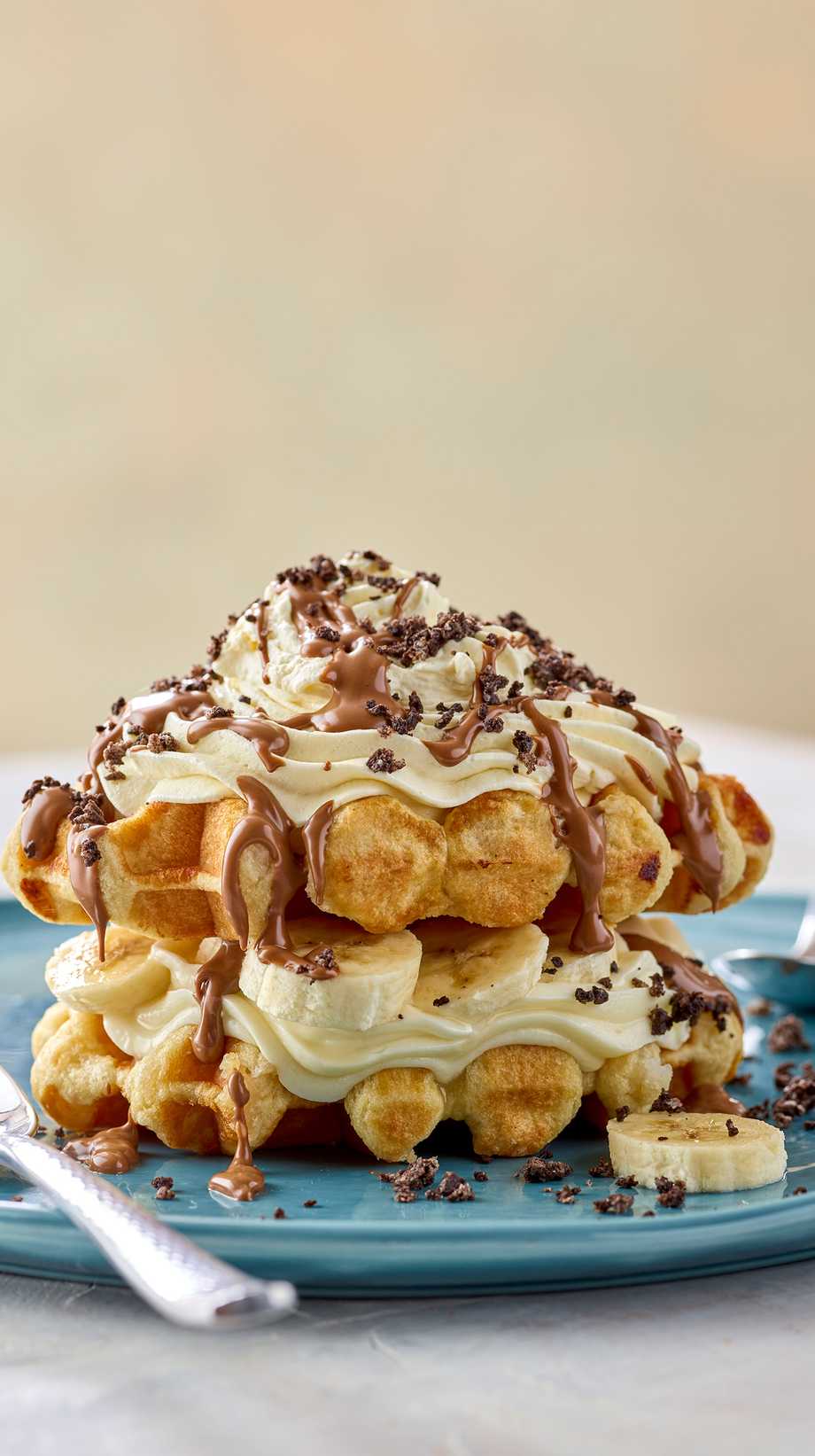 Waffles are covered with cream and chocolate on a blue plate. Two spoons are by its side.