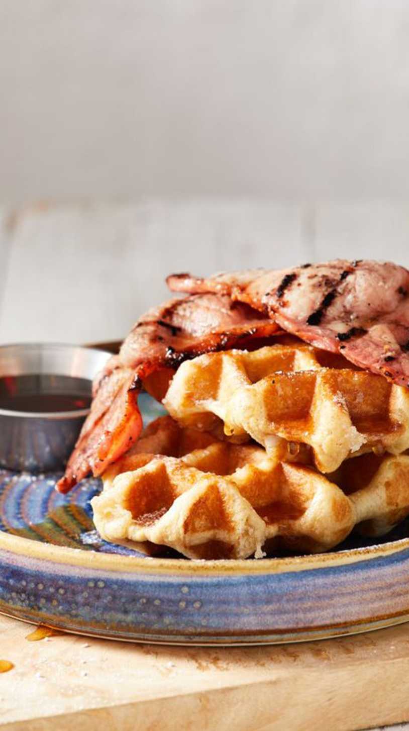 Crispy bacon sits on top of fresh waffles, with a tub of maple syrup nearby.