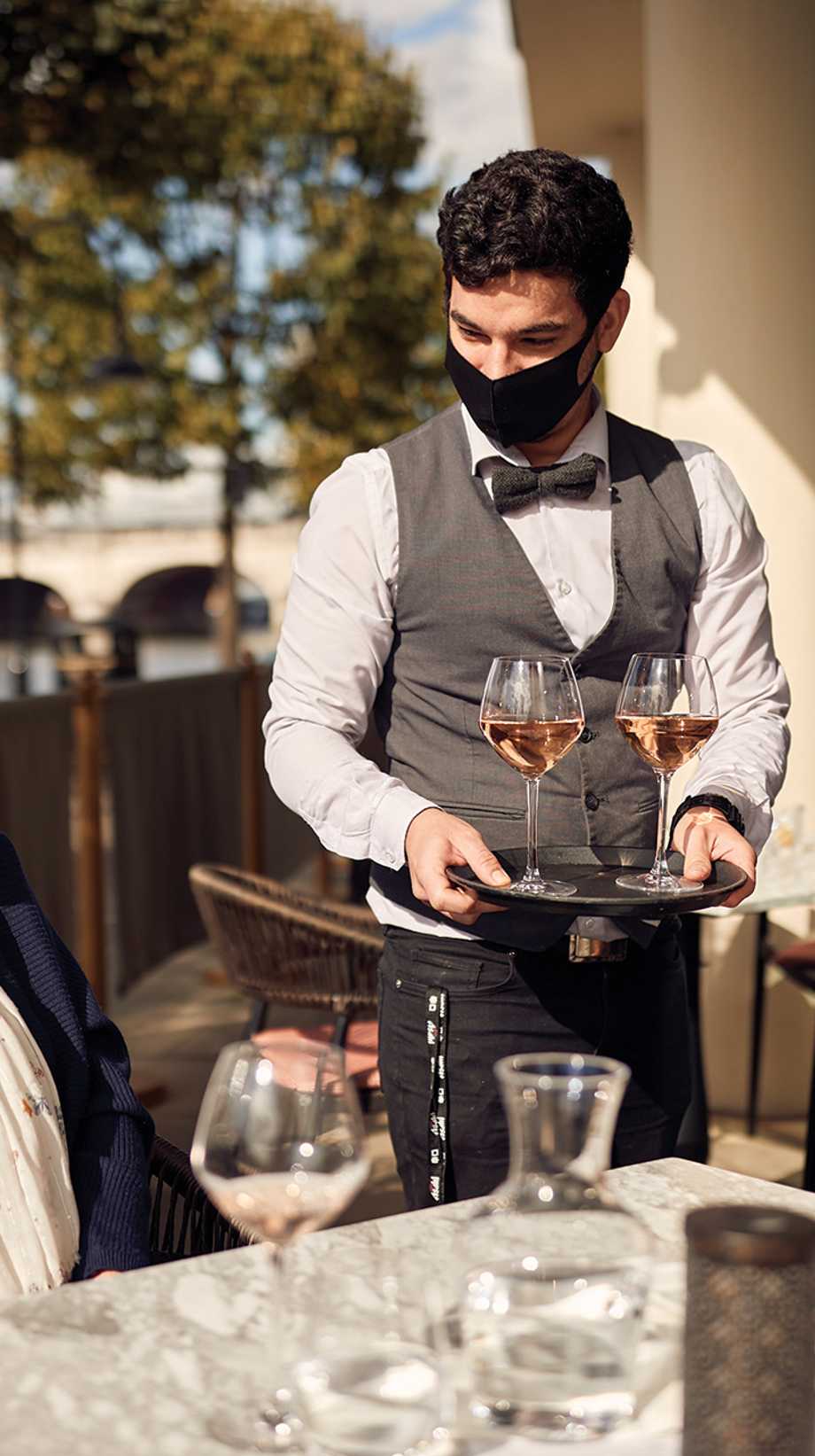 A team member wearing a face mask, waistcoat, and bowtie serves two glasses of wine to waiting guests, sitting in the sun.