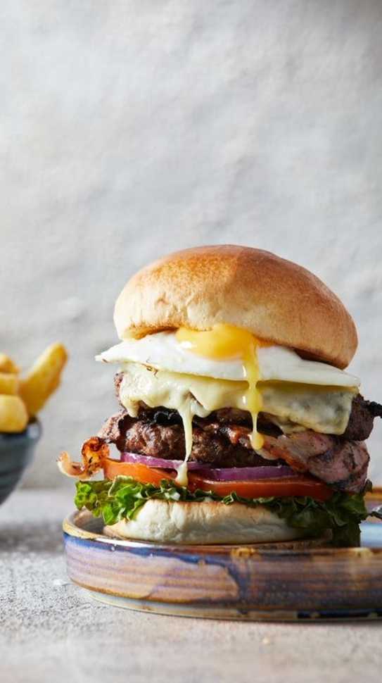 A towering burger sits on a plate. Underneath it's bun, golden yolk runs down.