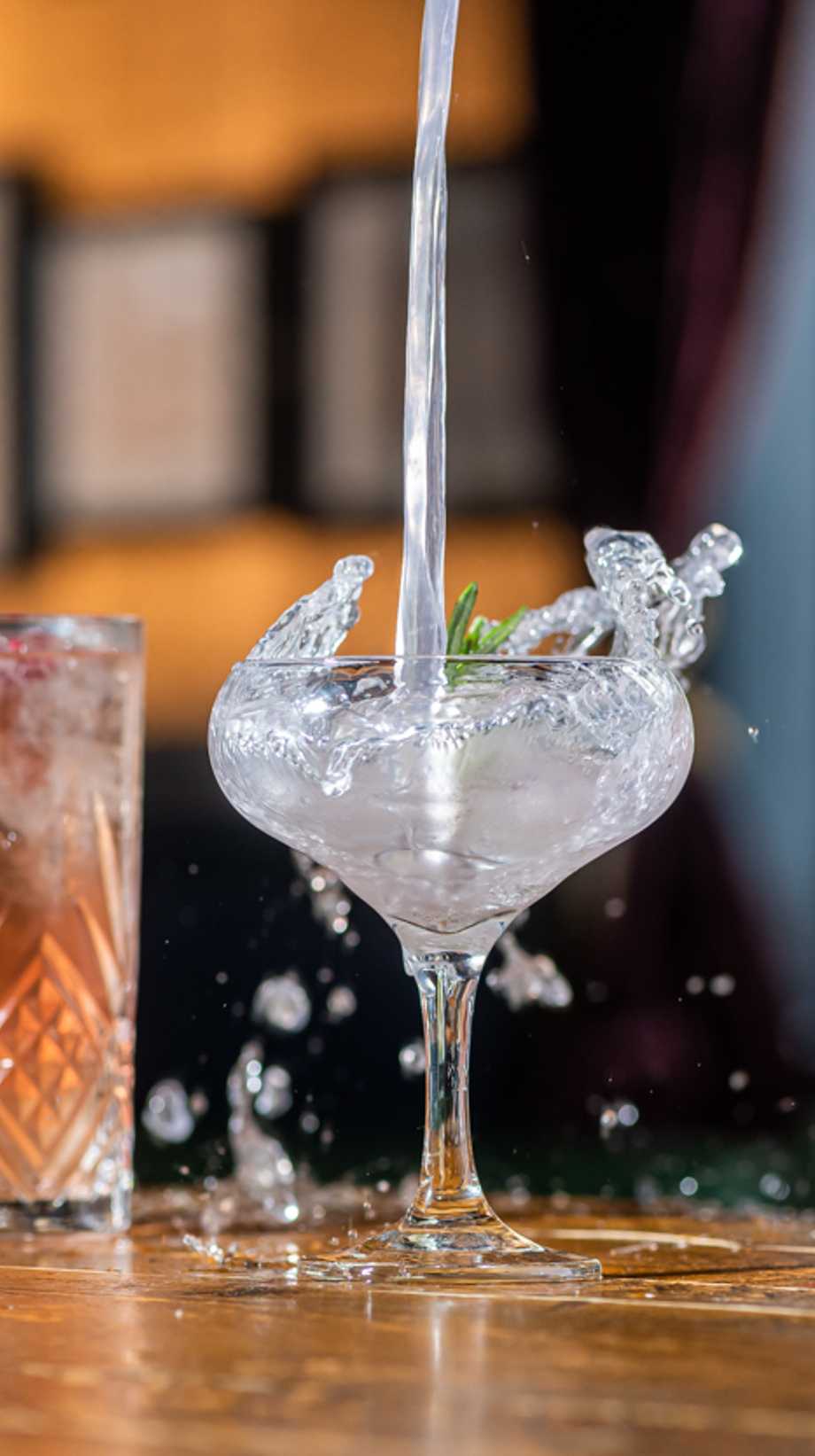 A cocktail is being poured from high above, creating a dramatic effect of ice and droplets outside the glass.