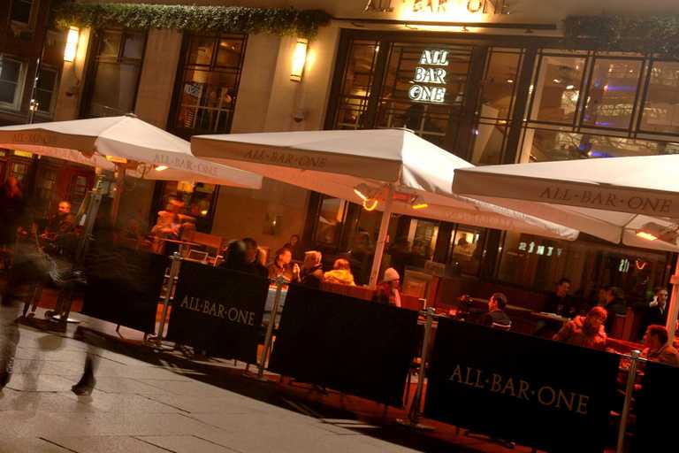 Tables and wide, white umbrellas are set up outside an All Bar One, where guests are sitting.