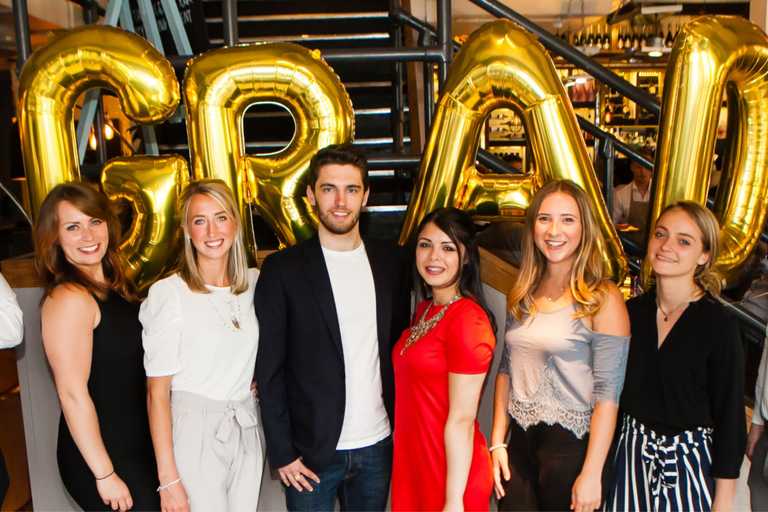 Five women and one man smile at the camera. Behind them, gold balloons spell out, "grad."