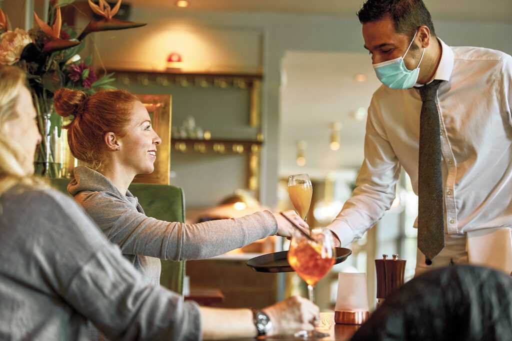 A team member wearing a face mask delivers a drink to a customer. Her friend sits in the foreground with an Aperol Spritz.