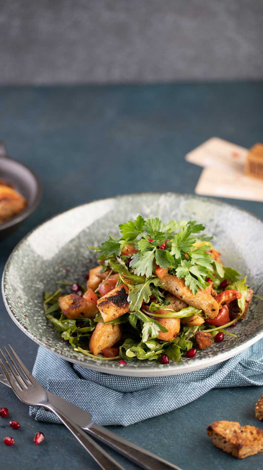 A fresh salad bowl sits on top of a blue serviette. The foreground has clean cutlery, pomegranate seeds, and crusty bread.