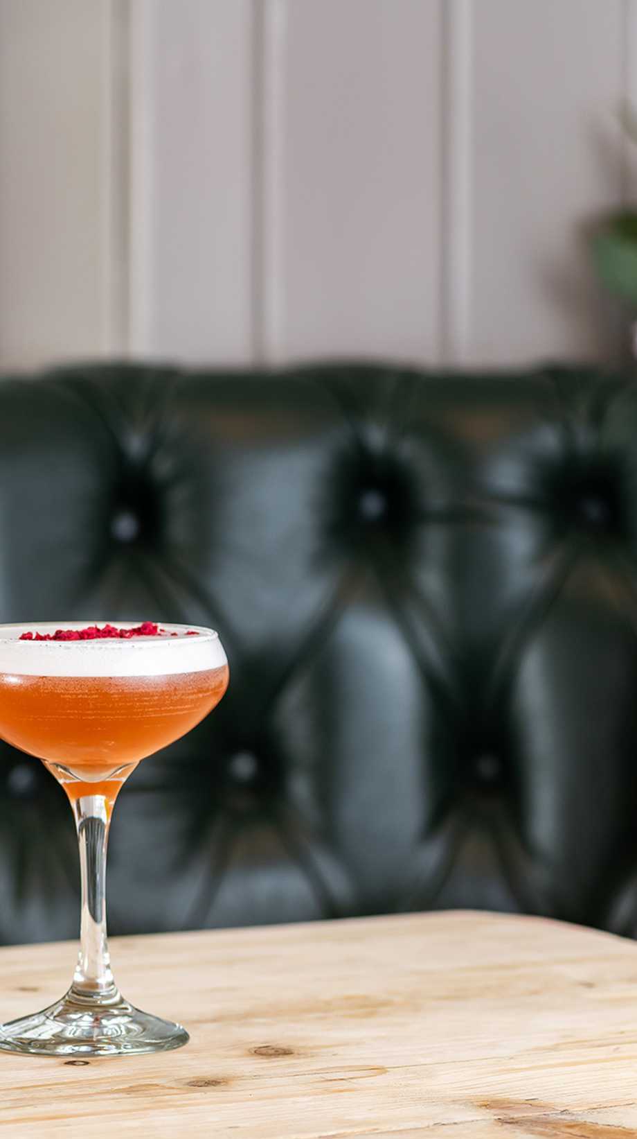 Two fruity cocktails are sitting on a table, set against a black leather booth and cushions.