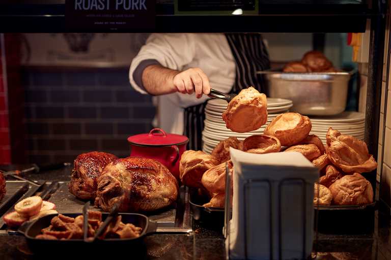 A chef piles fluffy Yorkshire puddings high on the carvery deck. Next to them are roasted meats and crispy parsnips.