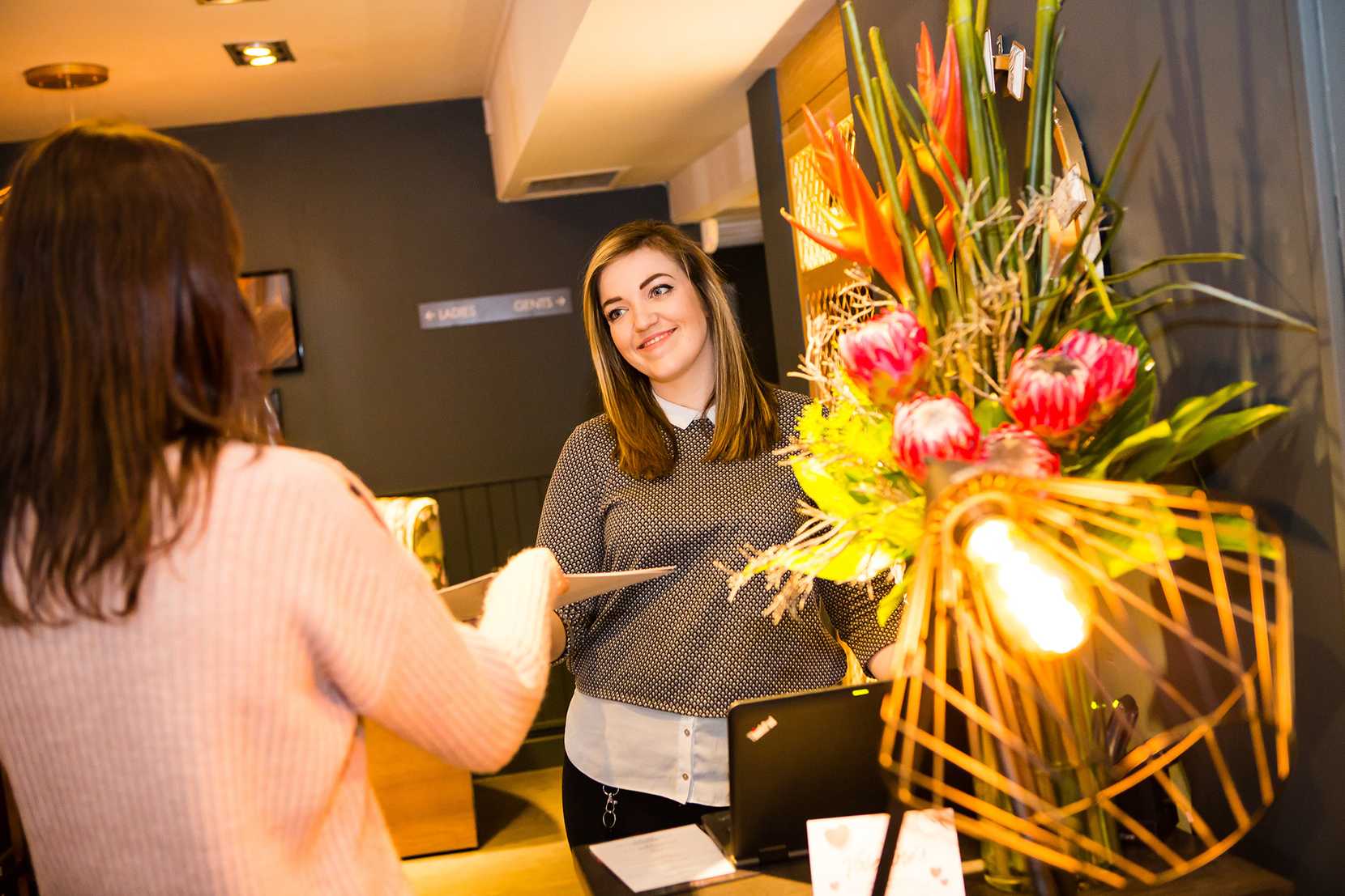 A guest is holding a menu. In the foreground, there's a modern lamp and flower display.