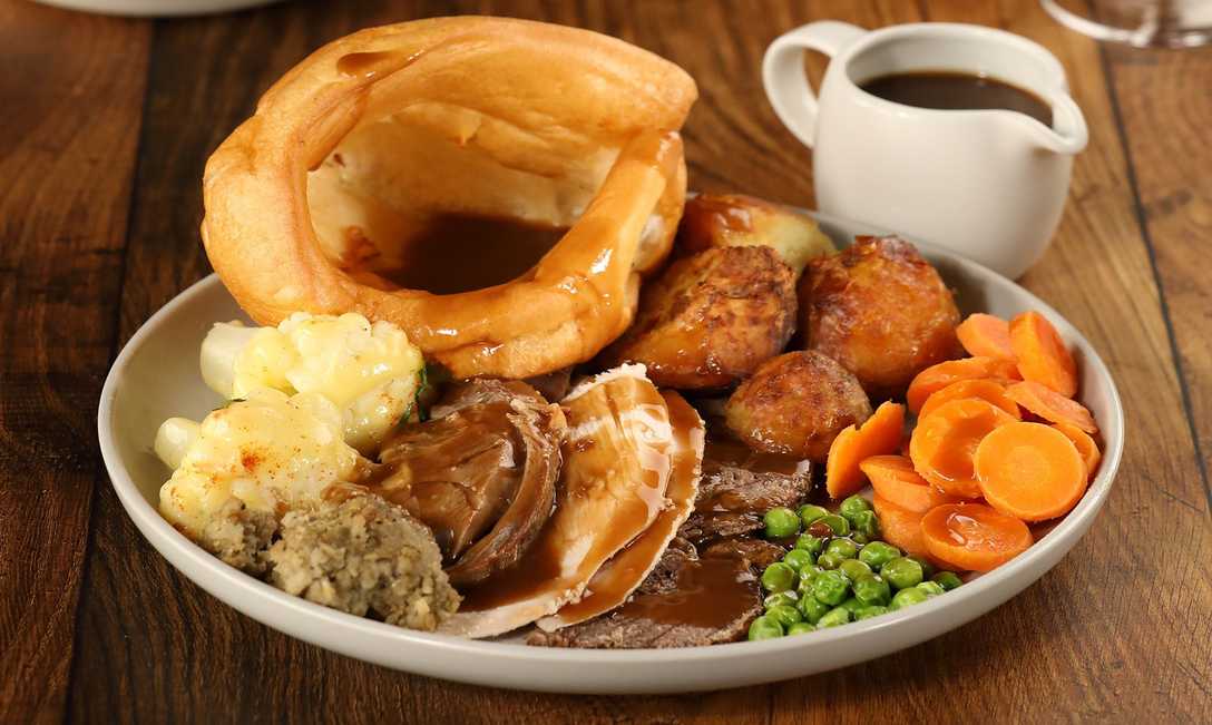 A close-up shot of a Toby Carvery roast, including cauliflower cheese, stuffing, carrots, peas, roast potatoes, a Yorkshire pudding, multiple meats, and a pot of gravy.