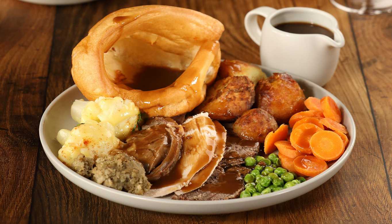 A close-up shot of a Toby Carvery roast, including cauliflower cheese, stuffing, carrots, peas, roast potatoes, a Yorkshire pudding, multiple meats, and a pot of gravy.