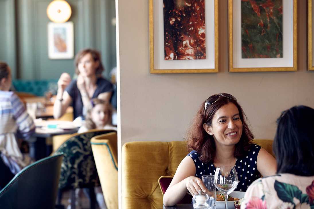 In the foreground, two guests laugh over glasses of wine. In the background, a family is sitting around a table.