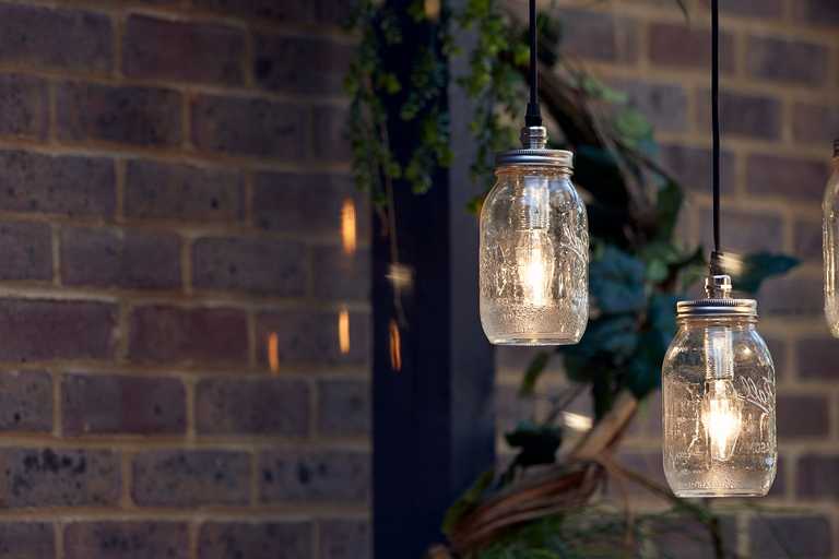 Glass jar pendant lights hang in front of a brick wall, plants, and beam, creating a cosy atmosphere.