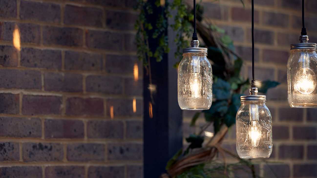 Glass jar pendant lights hang in front of a brick wall, plants, and beam, creating a cosy atmosphere.