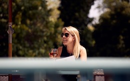 A woman wearing sunglasses is smiling at something off-camera, glass of wine in hand.