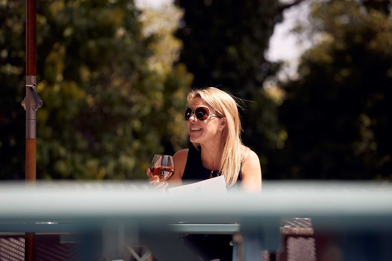 A woman wearing sunglasses is smiling at something off-camera, glass of wine in hand.