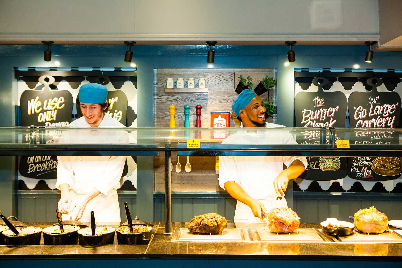 Two chefs are working at a Toby Carvery deck while smiling at someone off-camera.