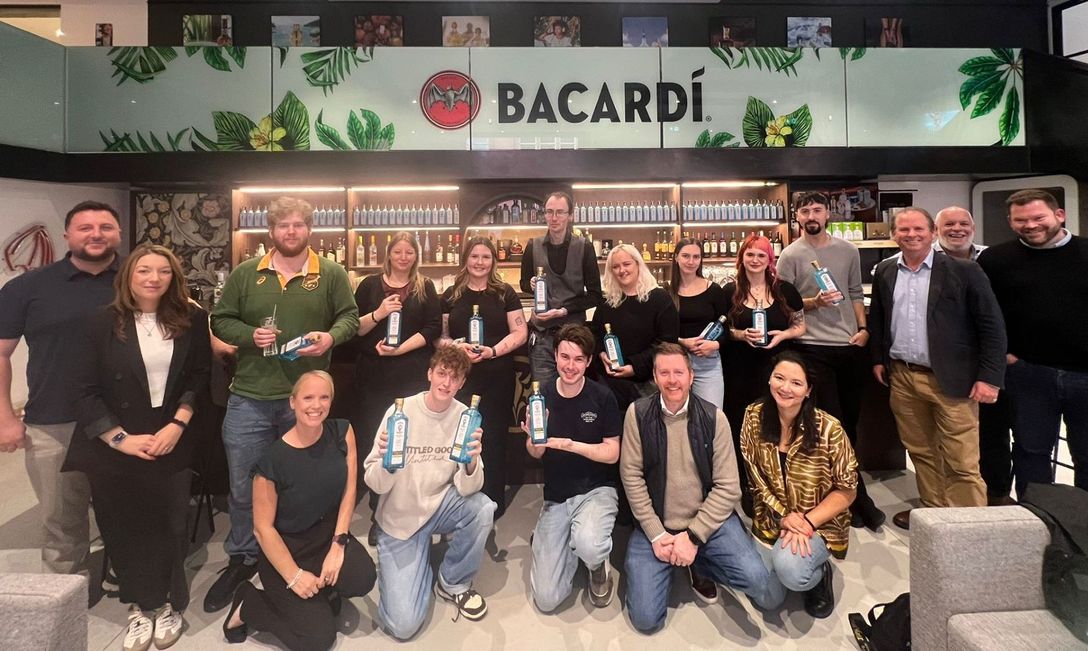 A group of Nicholson’s team members posing in front of a Bacardi-branded bar area, with shelves of bottles and green leaf decorations. Some individuals hold Bacardi bottles, capturing the celebratory spirit of the Annual Drinks Competition.