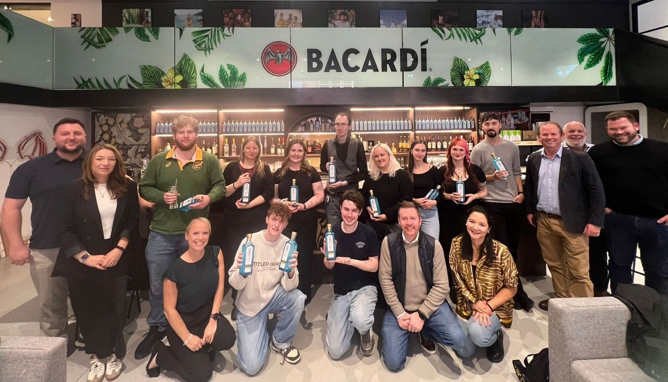 A group of Nicholson’s team members posing in front of a Bacardi-branded bar area, with shelves of bottles and green leaf decorations. Some individuals hold Bacardi bottles, capturing the celebratory spirit of the Annual Drinks Competition.