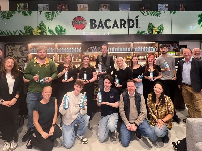 A group of Nicholson’s team members posing in front of a Bacardi-branded bar area, with shelves of bottles and green leaf decorations. Some individuals hold Bacardi bottles, capturing the celebratory spirit of the Annual Drinks Competition.