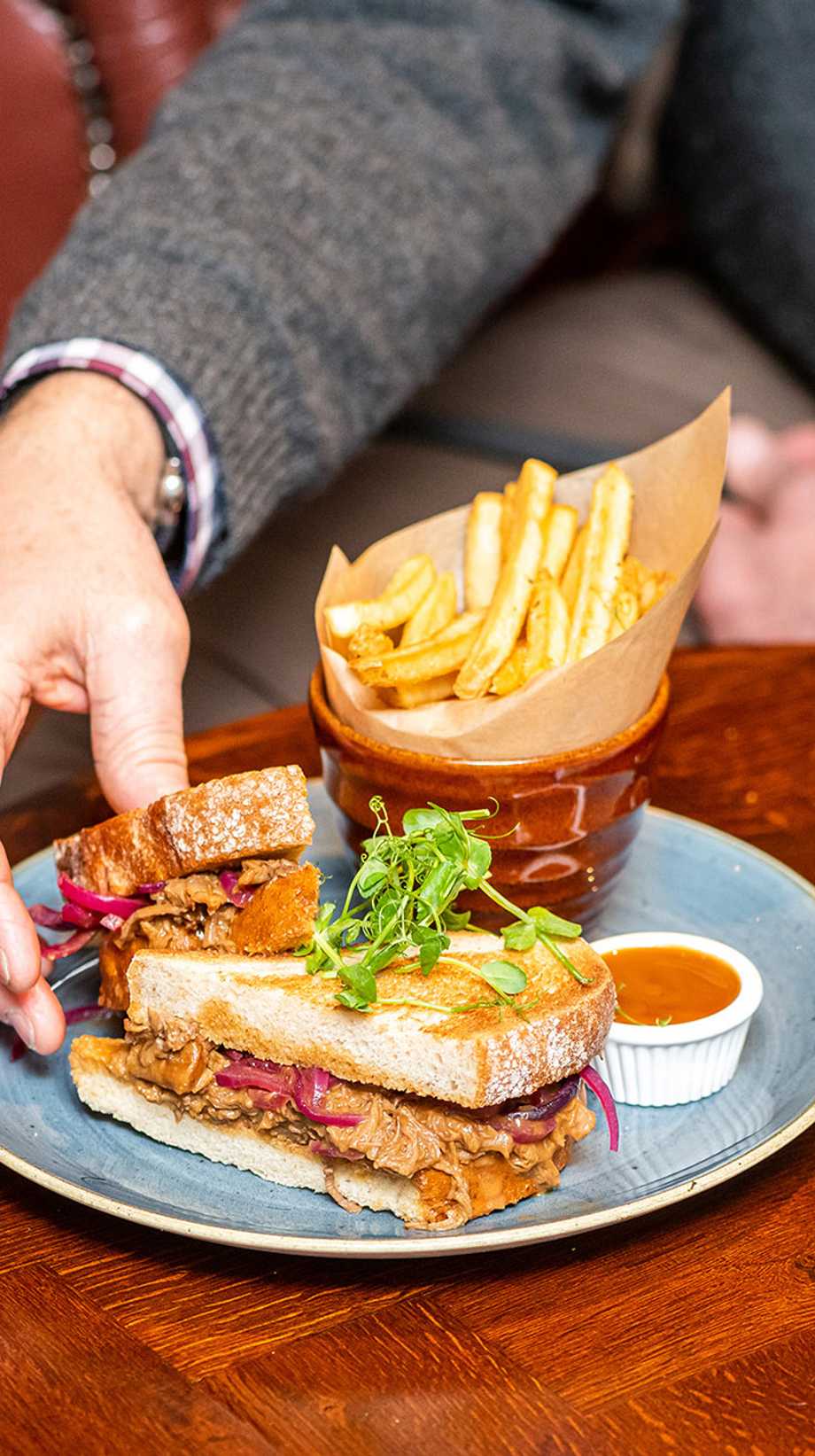 A man in a shirt and jumper reaches for half of his sandwich, resting next to a serving of fries and a pint.