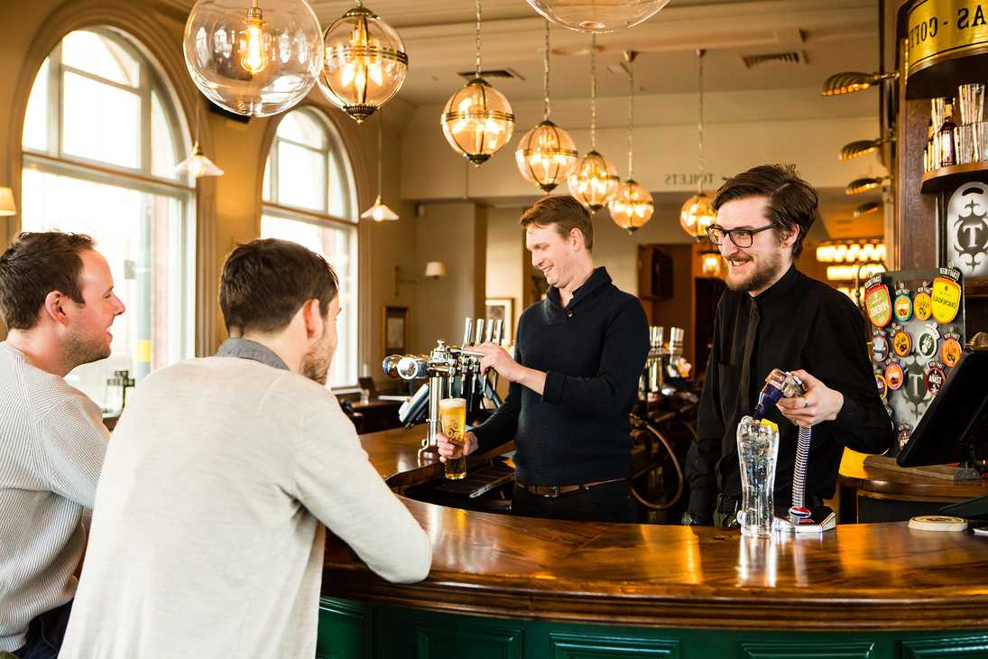 Two team members smile at guests while pouring a pint and a lemonade. Above them, pendant lights hang above the bar.