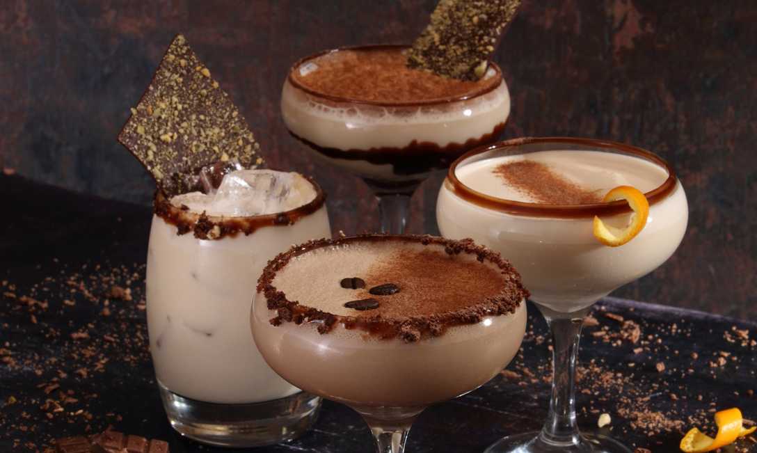 Four luxurious cocktails, each with a chocolate-related flavour. Each is garnished with something, from chocolate to coffee beans and orange peel. Chocolate is sprinkled around the glasses.