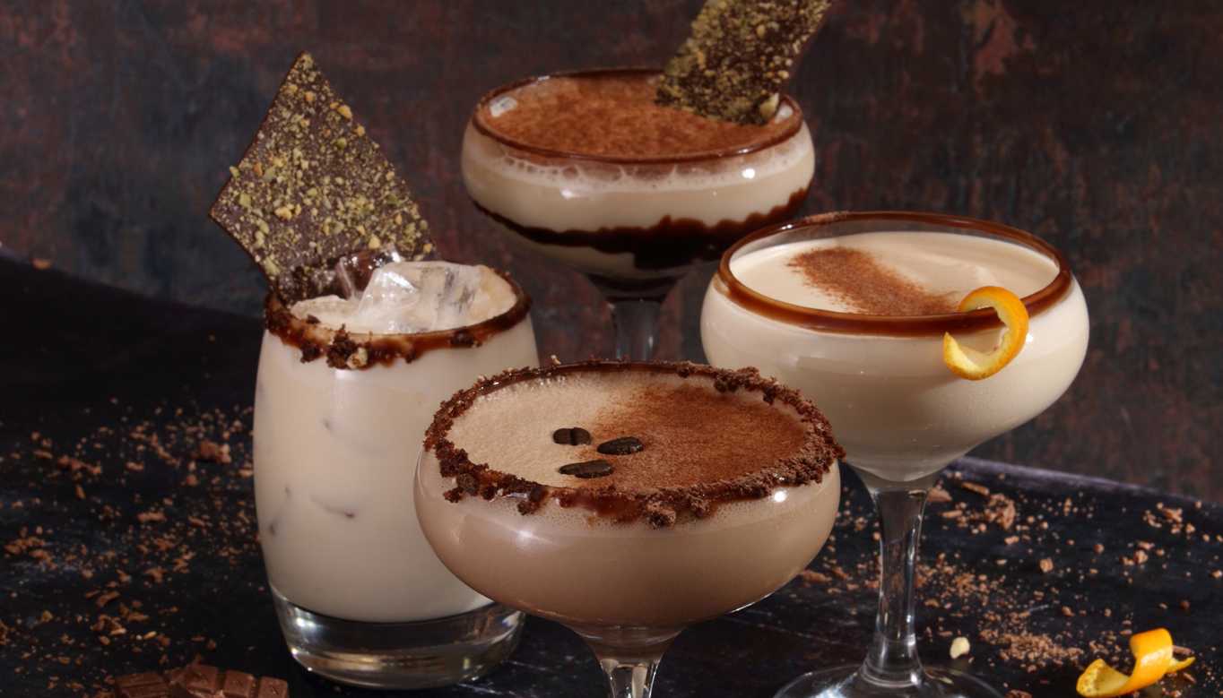 Four luxurious cocktails, each with a chocolate-related flavour. Each is garnished with something, from chocolate to coffee beans and orange peel. Chocolate is sprinkled around the glasses.