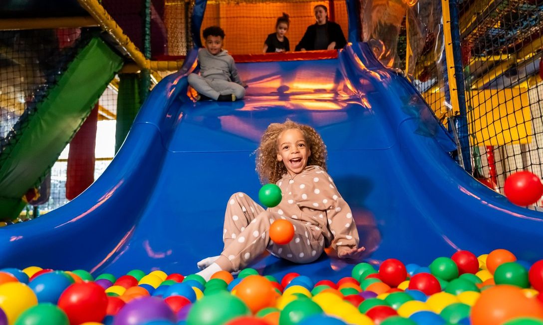 A child in a polka-dotted outfit slides down a blue slide into a colourful ball pit at an indoor play area, with two other children and an adult waiting at the top.