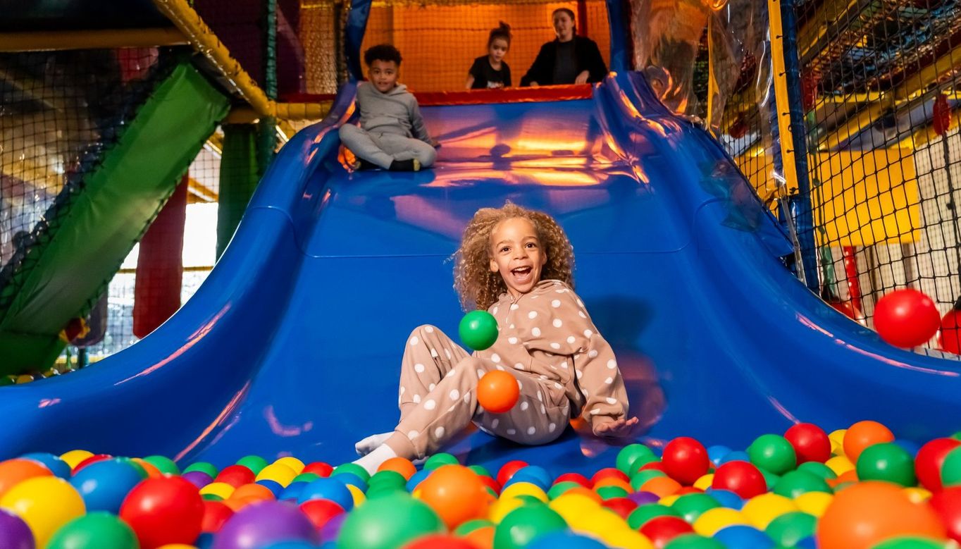 A child in a polka-dotted outfit slides down a blue slide into a colourful ball pit at an indoor play area, with two other children and an adult waiting at the top.