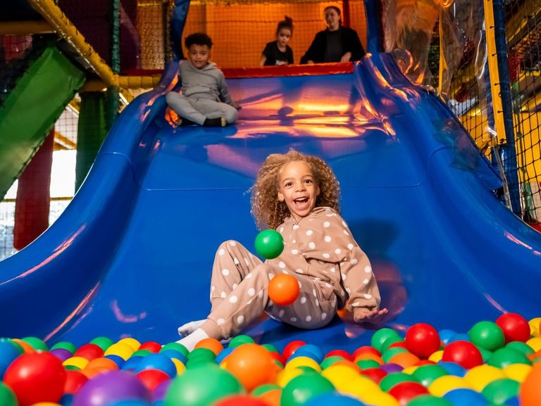 A child in a polka-dotted outfit slides down a blue slide into a colourful ball pit at an indoor play area, with two other children and an adult waiting at the top.