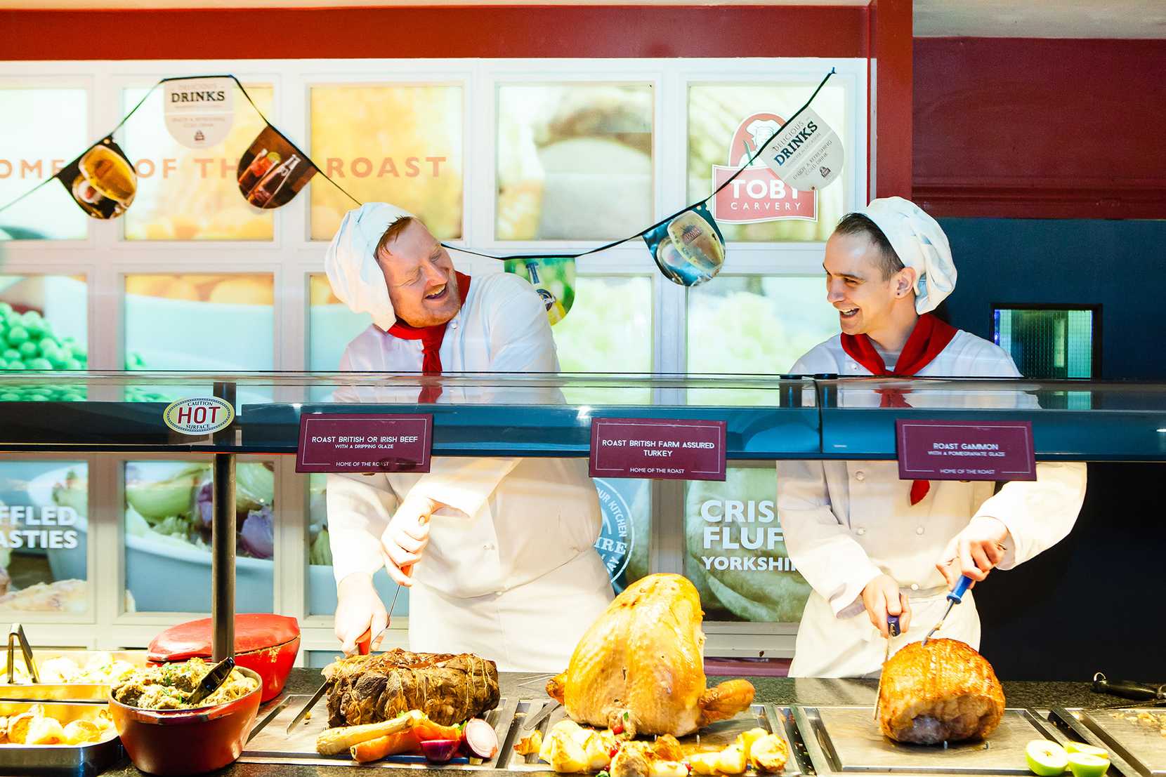 Two chefs are carving meat at a Toby Carvery deck while smiling.