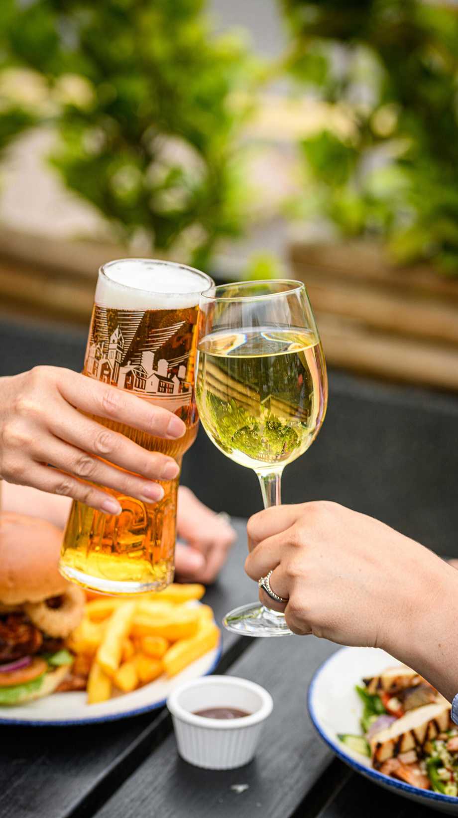 A guest with a beer raises a glass to another guest with a glass of wine. They are sitting outside on a sunny day.