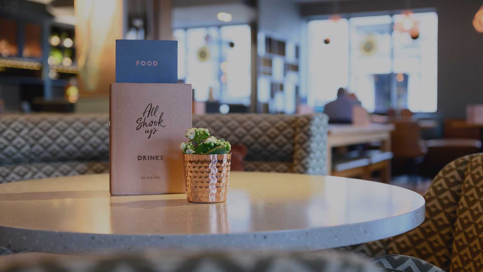 A menu reading, "All Shook Up" sits behind a copper plant pot with flowers inside.