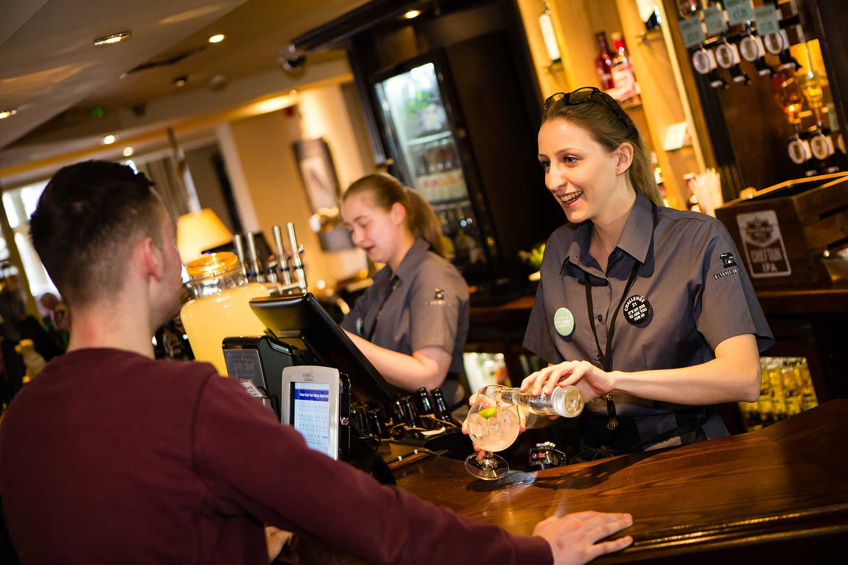 A team members creates a G&T for a guest while smiling. Behind them, another team member takes an order.