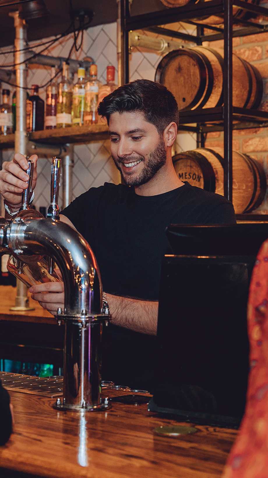 A bearded team member is pouring pints at the bar for waiting guests. Behind him, kegs are in front of an exposed brick wall.