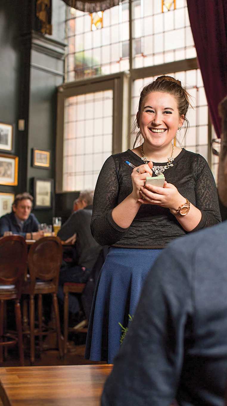 A team member takes a seated guests order. Behind them, a large window and more seated guests.