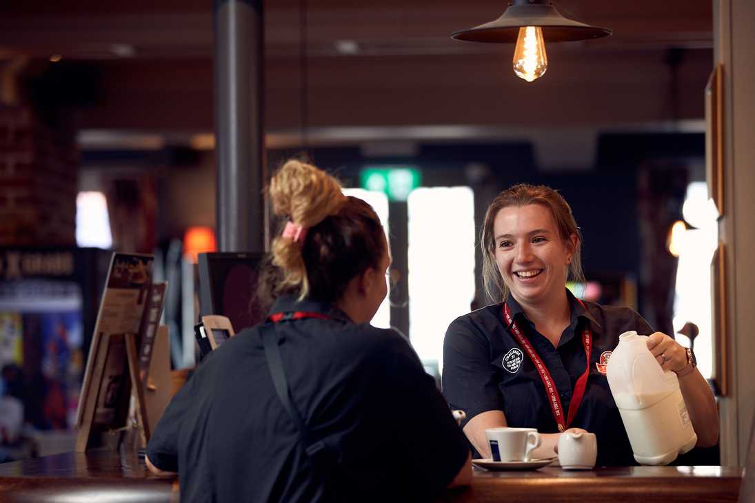 A team member prepares to pour milk into a small jug for a guest while chatting with another member of the team.