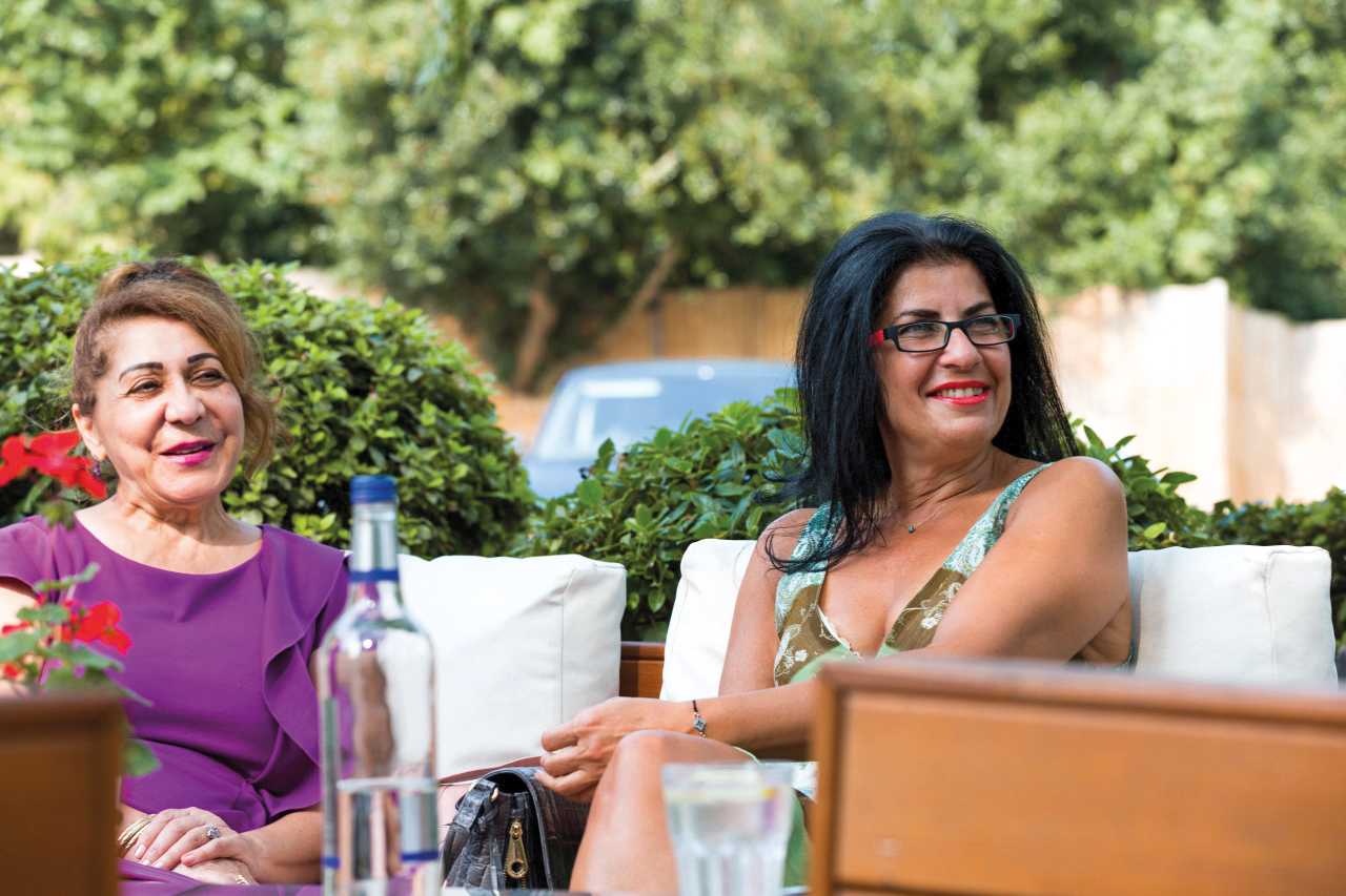 Two women sit outside on white cushions, speaking to someone off camera. In the foreground, there's a bottle of water and a red flower.