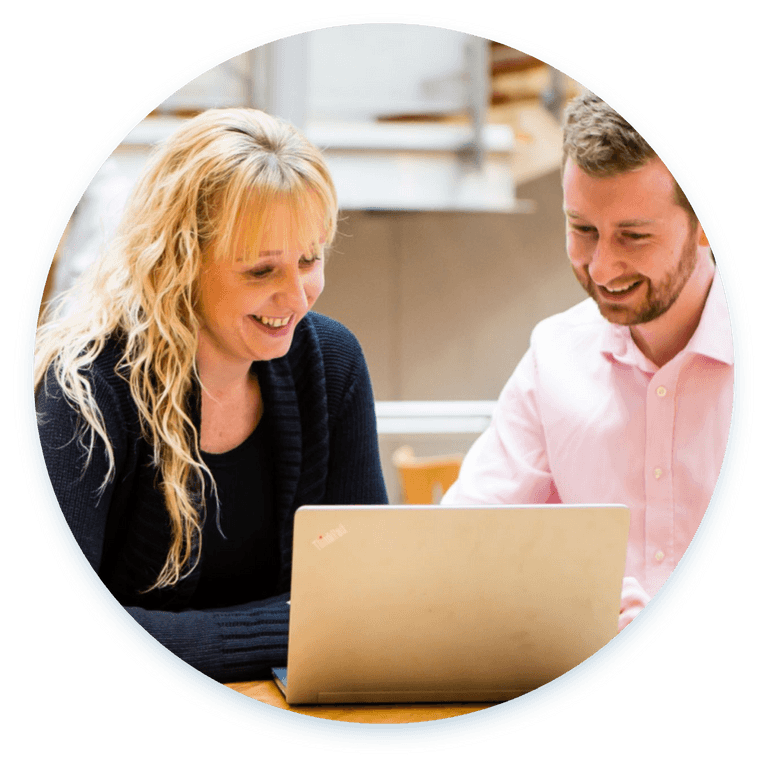 Two people are sitting at a table while discussing something on their laptop screen.