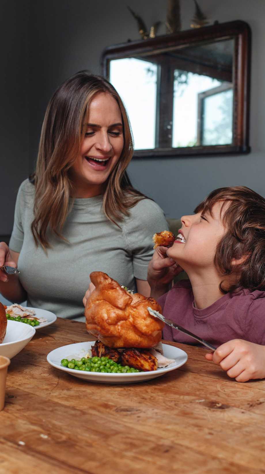 A mum feeds her son a piece of roast chicken from her force. The boy smiles, a large roast chicken and peas on his plate.