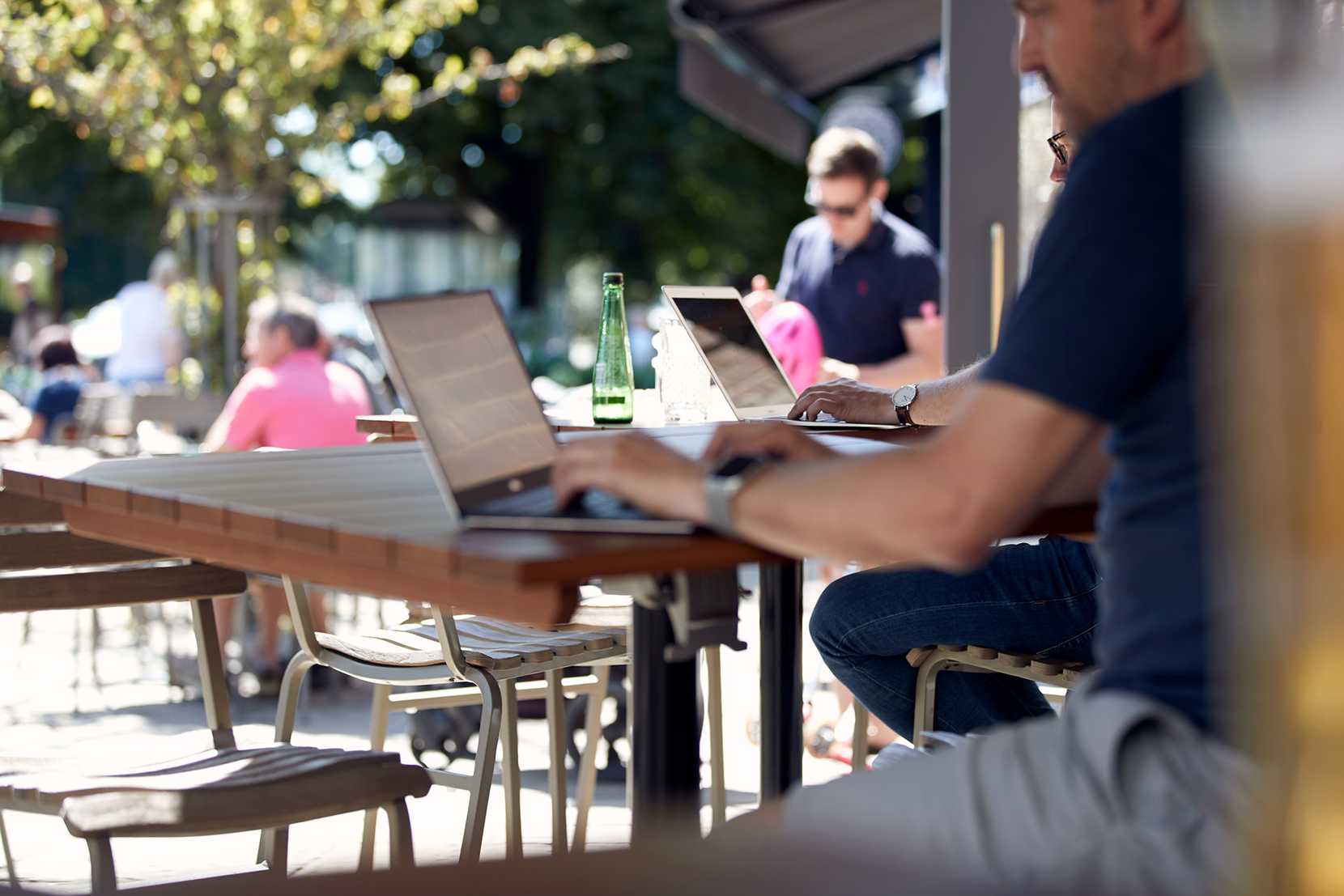 Two men are hard at work on their laptops, working while others enjoy a drink in the sun outside.