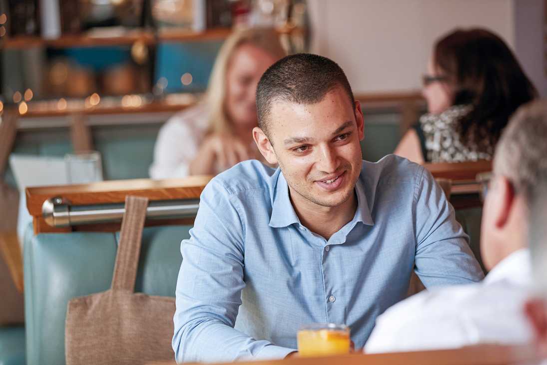 A man in a blue shirt is sitting in a booth with an orange juice, speaking to another guest.