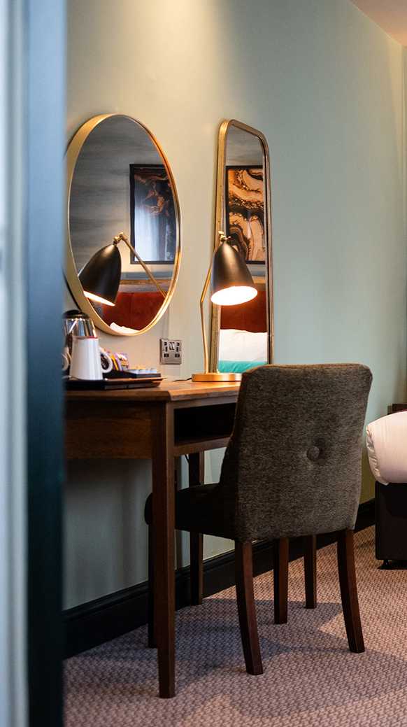 A gold-rimmed mirror sits above a wooden desk, where there's also a lamp and tea-and-coffee making area.