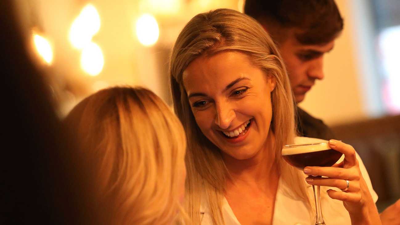 A lady with blonde hair smiles at a friend while holding a freshly made Espresso Martini. Behind them, the glow of warm lighting.