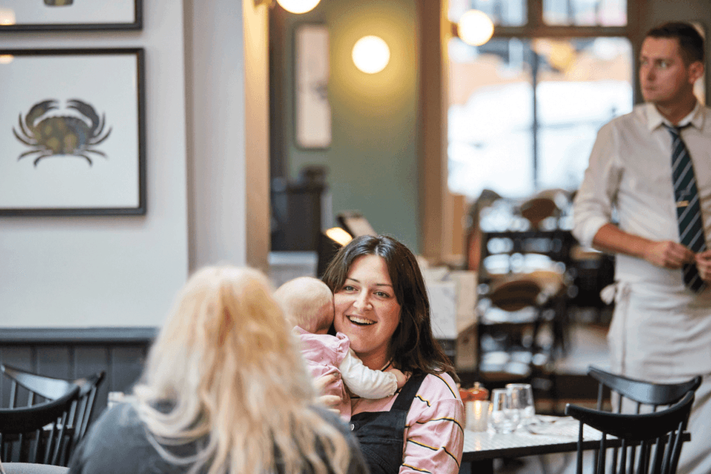 A guest is holding their new-born child to their chest while chatting with a friend. In the background, a team member walks past.