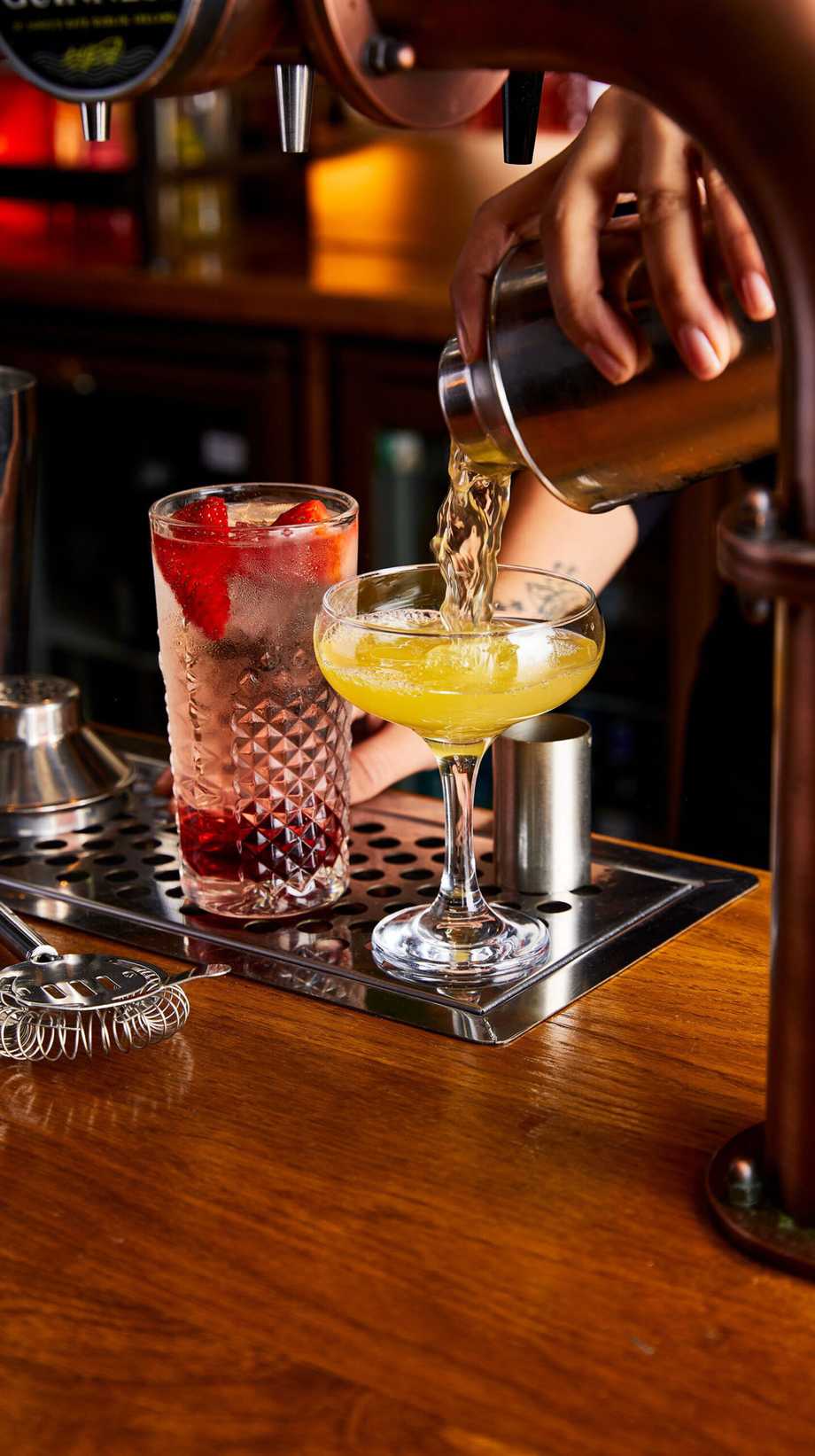 A team member is pouring a cocktail from a mixer into a glass. Next to it, a cocktail with a fruit garnish has been created.