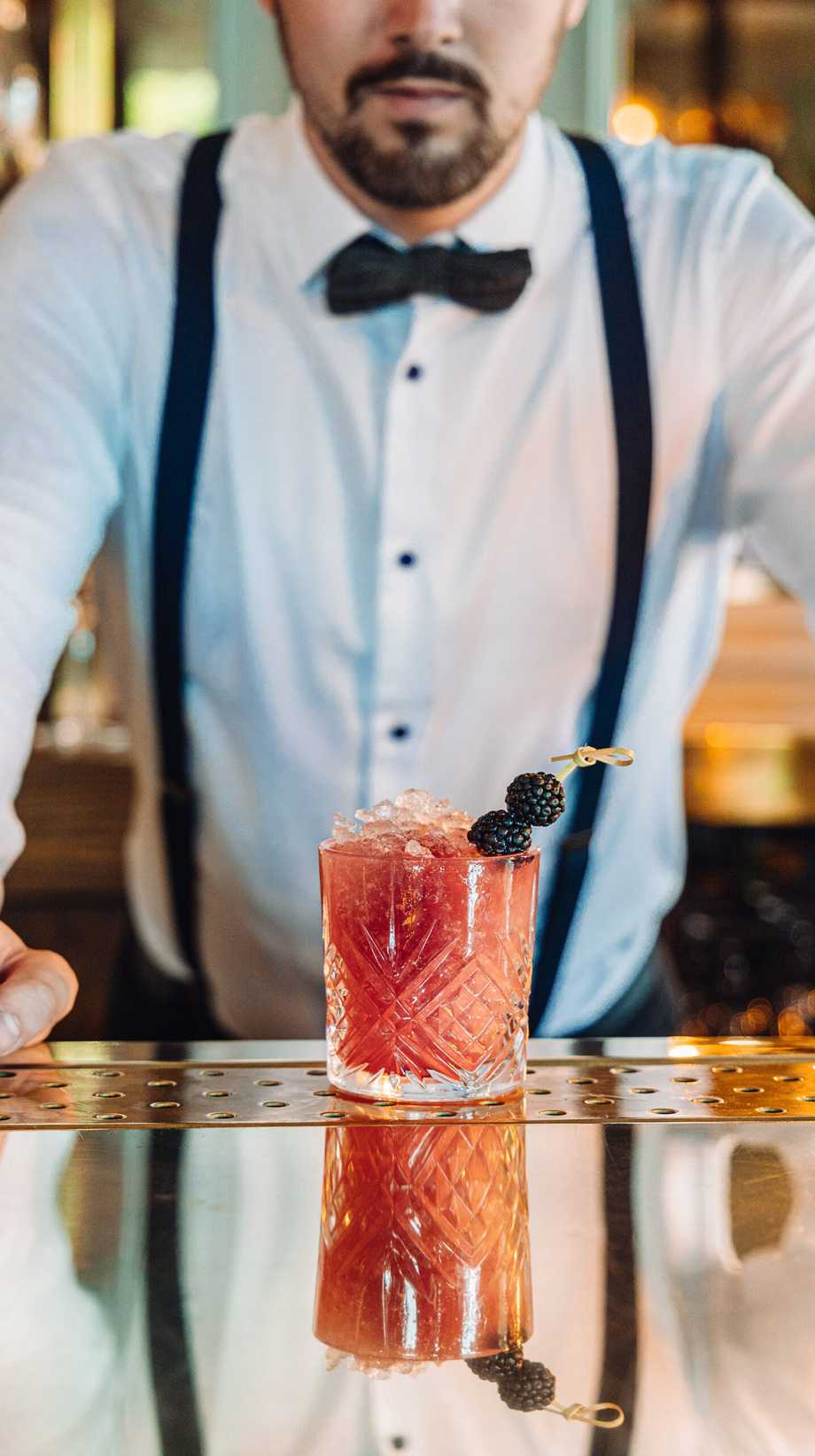 A mixologist wearing suspenders and a bow tie stands in front of a freshly-made cocktail.