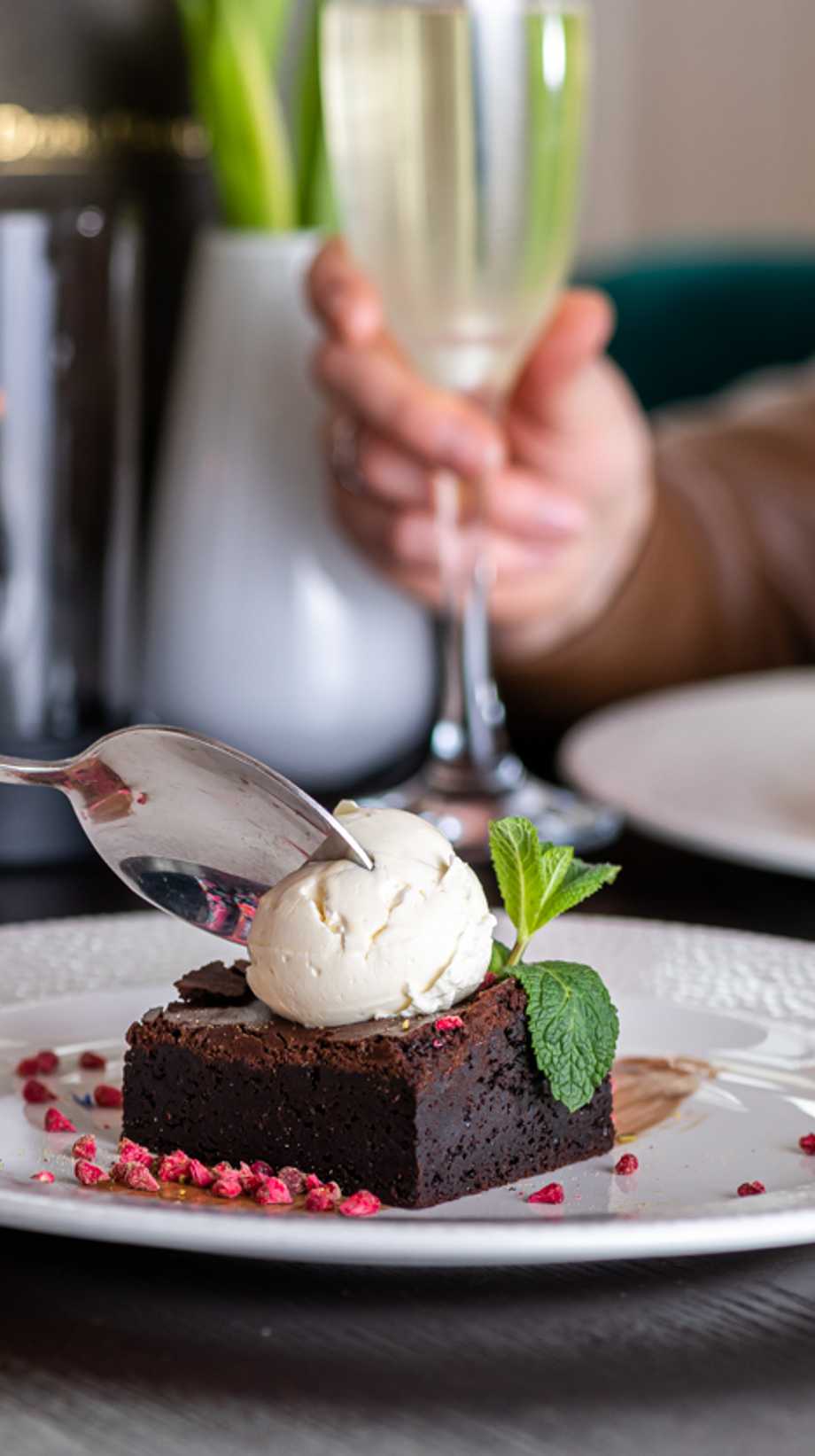 A guest is about to start eating a chocolate brownie with ice cream on top. In the background, a guest reaches for their drink.